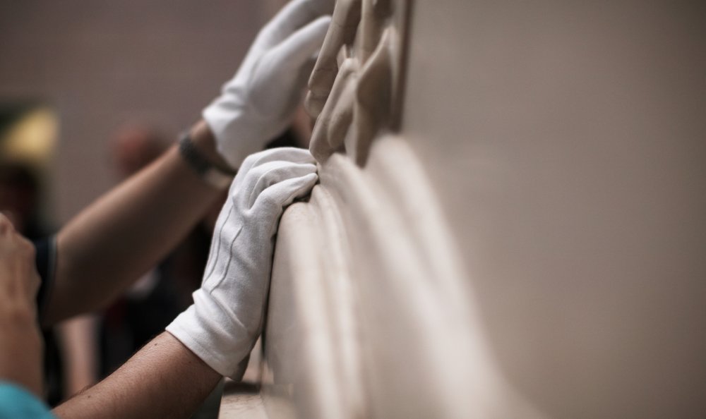 Close-up view a person's hands, with white cotton gloves on, touching a sculpture.