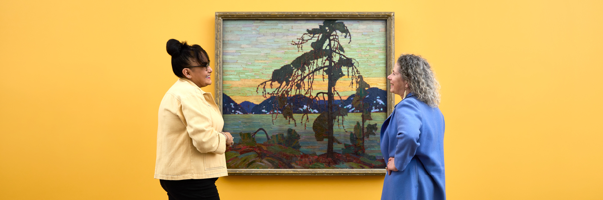 Medium shot of two individuals looking at Tom Thomson's The Jack Pine, hung on a yellow wall.