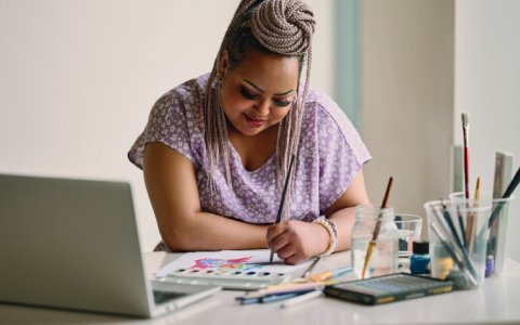 A woman painting with watercolours in front of her laptop.